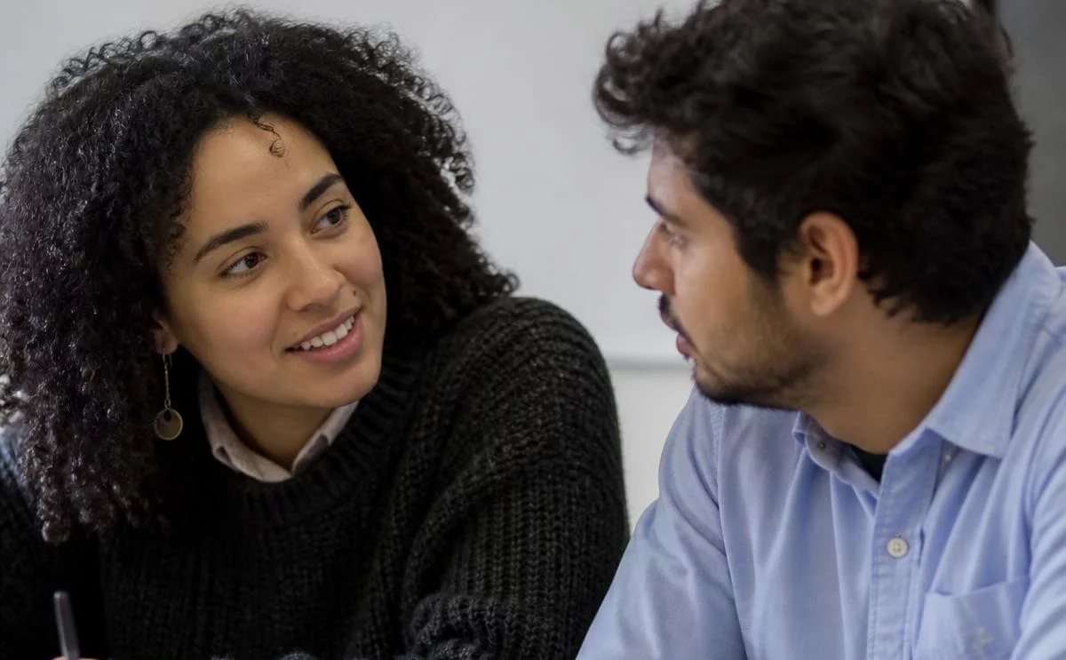 Duas pessoas frente a frente na mesa: uma sustenta a fala, a outra anota o que ouviu